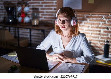 Young Redhead Woman Working At The Office At Night Wearing Headphones Relaxed With Serious Expression On Face. Simple And Natural Looking At The Camera. 