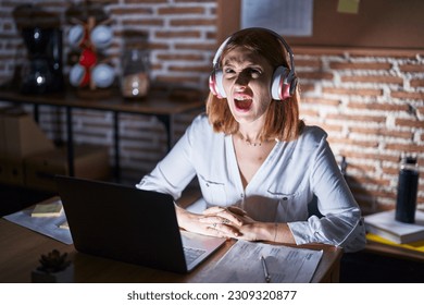 Young redhead woman working at the office at night wearing headphones angry and mad screaming frustrated and furious, shouting with anger looking up.  - Shutterstock ID 2309320877