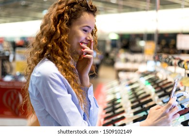 Young Redheaded Girl Standing At The Counter In An Electronics Store Choosing A New Phone To Buy Taking A Selfie Checking The Camera