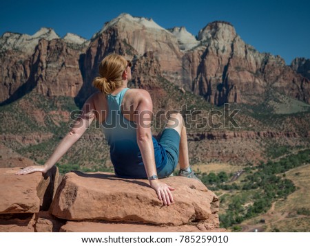 Young redheaded sportswoman reclines in the hot sun on a red rock outcrop in Zion National Park. Red and white canyon walls frame the background against a blue sky. 