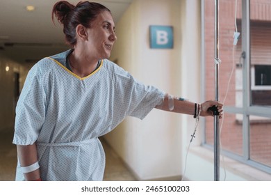 Young redheaded woman post-surgery, walking with IV drip in hospital gown, looking out hospital window. - Powered by Shutterstock