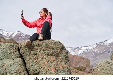 Young Redheaded Woman With Red Jacket Sitting On A Rock In The Middle Of The Andes Mountains Of Chile, Taking A Picture With Her Phone