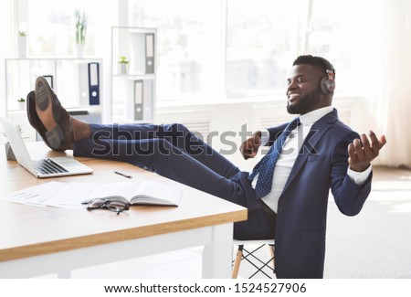 Young relaxed african businessman having fun at office, listening to music and perfoming as playing guitar, putting legs on workdesk, copy space