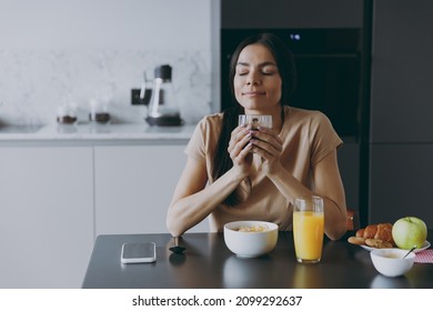 Young Relaxed Brunette Housewife Caucasian Woman 20s In Casual Clothes Beige Shirt Eat Breakfast Drink Coffee Sniff Scent Cooking Food In Light Kitchen At Home Alone. Healthy Diet Lifestyle Concept