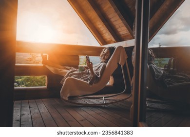 Young relaxed cheerful woman enjoying nature on the balcony of the cottage at sunset with a cup of tea and a smartphone in her hands - Powered by Shutterstock