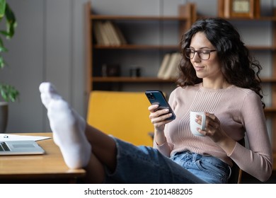 Young relaxed female employee holding smartphone and tea cup resting at workplace, selective focus. Woman relaxing on chair with legs on office table, scrolling social media while taking break at work