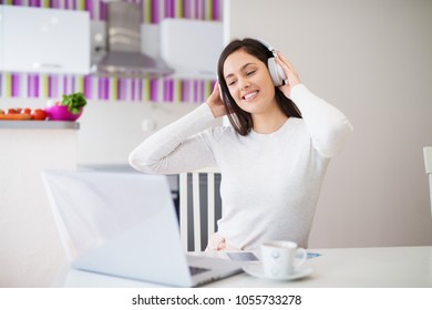 Young Relaxed Happy Girl With Headphones On Looking At Laptop Drinking Coffee And Enjoying Music While Sitting At The Kitchen Table.