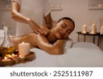 Young relaxed lady enjoy body massage lying on table at spa salon with closed eyes, masseur rubbing her shoulders and back in warm candles atmosphere