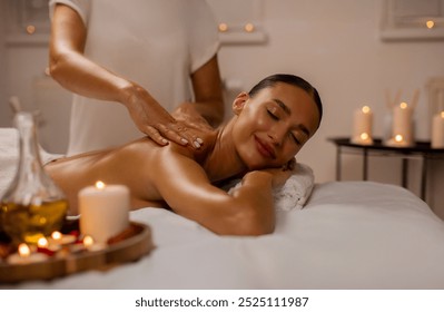 Young relaxed lady enjoy body massage lying on table at spa salon with closed eyes, masseur rubbing her shoulders and back in warm candles atmosphere - Powered by Shutterstock