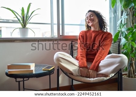 Young relaxed smiling pretty woman relaxing sitting on chair at home. Happy positive beautiful lady feeling joy enjoying wellbeing and lounge chilling near window in modern cozy apartment interior.