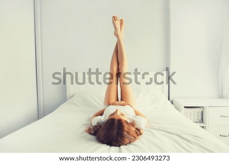Young relaxed woman lifts her legs up lying on the bed on soft mattress in white bedroom