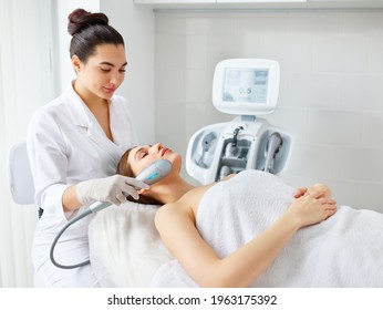 Young relaxed woman receiving professional facial treatment and SMAS lifting with laser machine from cosmetologist in modern beauty salon