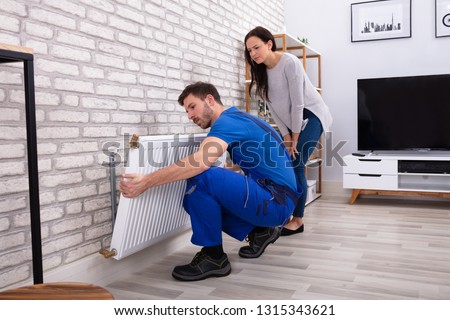Young Repairman Installing Radiator On Brick Wall With Woman Standing At Home