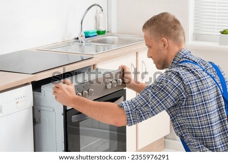 Young Repairman In Overall Repairing Oven In Kitchen