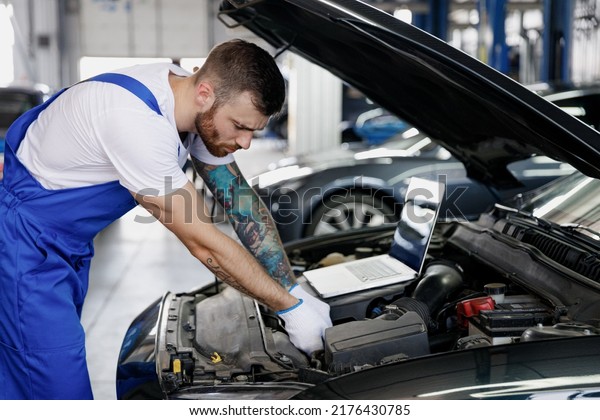 Young repairman professional technician car\
mechanic man in blue overalls t-shirt using laptop pc computer make\
diagnostics fix problem with raised hood work in vehicle repair\
shop workshop indoor.