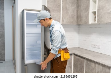 Young Repairman With Tool Belt Looking At Broken Refrigerator