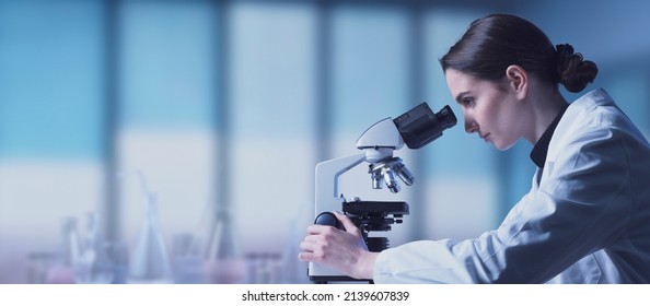 Young researcher working in the lab, she is looking through a microscope and checking samples, medical research concept