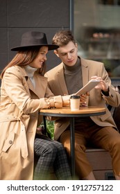 Young Restful Couple Sitting By Small Round Table Against Modern Architecture, Having Drinks And Discussing Online News While Using Tablet