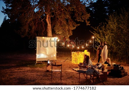 Young restful friends having drinks and watching movie on white screen in the evening in natural environment while sitting on the ground