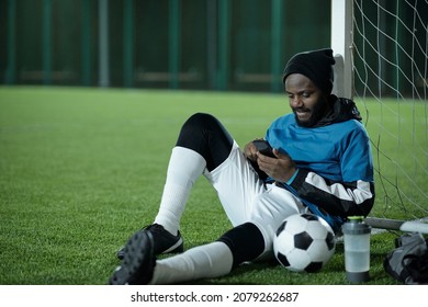 Young restful sportsman sitting by net and scrolling in smartphone while looking through news or online videos - Powered by Shutterstock