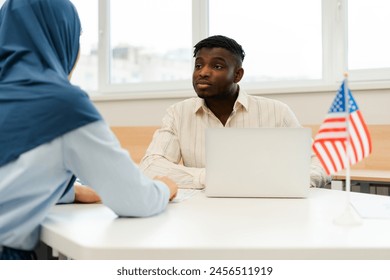 Young, retired African American male worker talking to female voter wearing hijab while sitting at registration table at polling station. Concept of presidential elections, democracy, freedom - Powered by Shutterstock