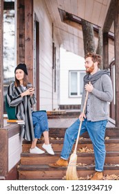 Young Rich Couple Spend A Quiet Peaceful Day Outdoor In Their Country House, Relaxing From The Urban Fuss. Young Man Sweeps The Leaves, Clean The Garden. Private House Purchase Concept.