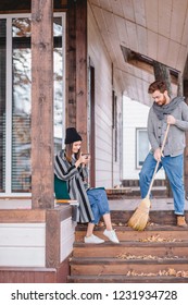 Young Rich Couple Spend A Quiet Peaceful Day Outdoor In Their Country House, Relaxing From The Urban Fuss. Young Man Sweeps The Leaves, Clean The Garden. Private House Purchase Concept.