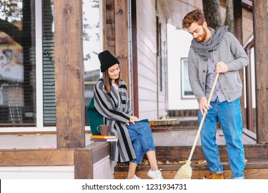 Young Rich Couple Spend A Quiet Peaceful Day Outdoor In Their Country House, Relaxing From The Urban Fuss. Young Man Sweeps The Leaves, Clean The Garden. Private House Purchase Concept.