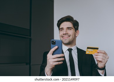 Young Rich Employee Business Man Corporate Lawyer In Black Suit Shirt Tie Read Document Using Mobile Cell Phone Credit Bank Card Shopping Online Order Delivery Booking Tour Sit At Table In Kitchen.