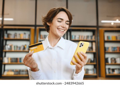 Young rich successful employee business woman wear white shirt casual clothes use mobile cell phone hold credit bank card buy online sit work at office desk with pc laptop. Achievement career concept - Powered by Shutterstock