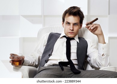 Young rich successful man sitting in white leather armchair with cigar and glass of whiskey. Fashion style portrait in modern interior.