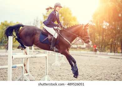 Young rider girl performing jump at horse show jumping competition. Equestrian sport background. Warm color toned image