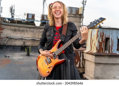 Young rock blonde girl with tattoos and two funny ponytails, laughing loudly and playing an electric guitar on the roof of an old building, front view. Musical video clip shooting. Outdoor show. - Shutterstock ID 2043985646
