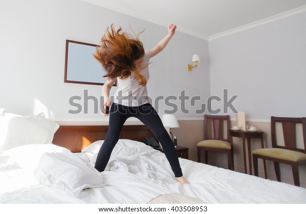 Young rock music fan girl expressively jumping on\
the bed in bright bedroom