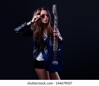 Young Rockstar Woman With Blue Electric Guitar Wearing Sunglasses Over Dark Background