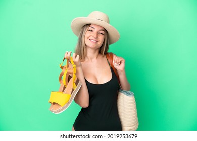 Young Romanian woman in swimsuit holding sandals for go to the beach isolated on green background smiling a lot