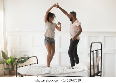 Young romantic african american couple wearing pajamas dancing on bed at home, happy active mixed race husband and wife enjoying funny party on honeymoon together in the morning in bedroom interior - Powered by Shutterstock