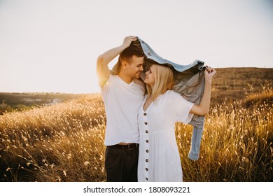 Young Romantic Couple In A Field With Dry Golden Grass, Hugging, Hiding Under A Jacket.