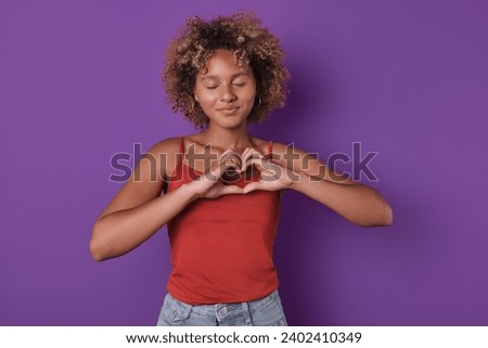Young romantic cute African American woman making heart shape from fingers and closing eyes smiling confessing love to boyfriend and posing for romantic valentine day card stands on purple background.