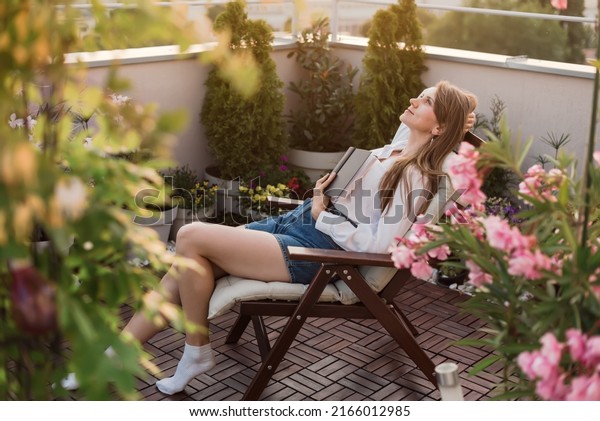 Young romantic woman holding book to her chest on\
urban rooftop garden at sunset. Dreamy girl reading exciting novel\
while sitting in chaise longue on cozy terrace with flowers, plants\
and city view.