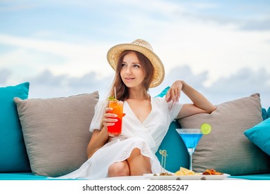 Young romantic woman in straw hat holding mai tai sits by table with alcoholic blue lagoon cocktail in martini glass at outdoors bar, restaurant at luxury resort, hotel. Relax, rest, holiday, vacation