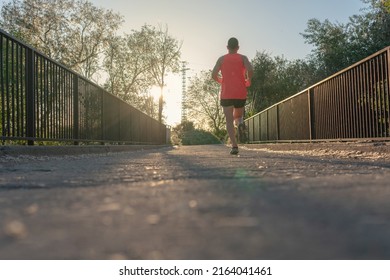 Young Runner Man With Orange T-shirt On The Asphalt Of The River Bridge. Agile Athlete Who Is Doing A Continuous Race With The Sunset In The Background Wearing The Shoes That Match The Shirt.