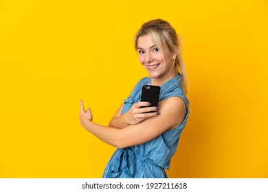 Young Russian Woman Isolated On Yellow Background Using Mobile Phone And Pointing Back