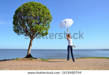 A young Russian woman in a white top and blue jeans standing near a round crown tree and with a white sun umbrella raised high in her hand. Cloudless sky and blue water background, sand upfront.