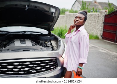 Young sad african woman stand with emergency triangle against car with popped hood. Transportation, vehicles problems and breakdowns concept.