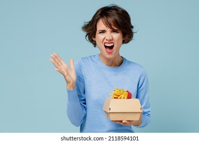 Young Sad Angry Woman Wearing Sweater Hold A Box With Fastfood French Fries Potato Burger Scream Shout Spread Hand Isolated On Plain Pastel Light Blue Background. People Lifestyle Junk Food Concept