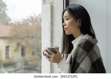 Young Sad Asian Woman Drinks Coffee Or Tea And Looks Out The Window Covered With A Blanket