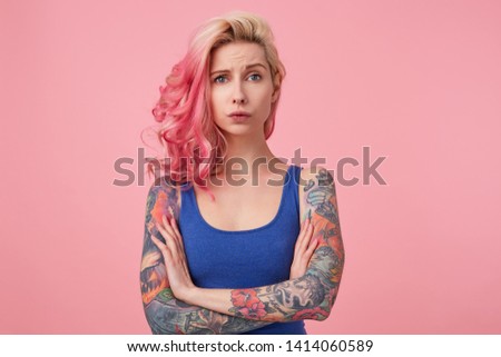 Young sad beauty woman with pink hair, stands with crossed arms over pink background, looks displeased and unhappy, wears a blue shirt. People and emoyion concept.
