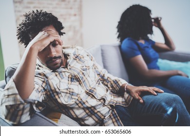 Young Sad Black Couple.Upset Man Being Ignored By Partner At Home In The Living Room.American African Men Arguing With His Stylish Girlfriend,who Is Sitting On Sofa Couch Next To Him.Blurred.