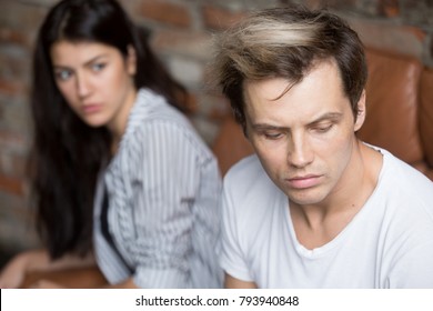 Young sad cheated man thinking of betrayal unresolved problem in bad relationship, upset girlfriend at background, depressed husband sitting his back to frustrated wife, unhappy couple after quarrel